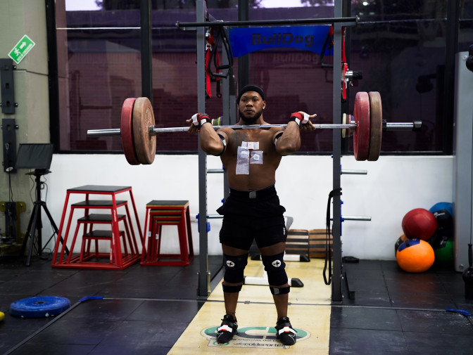 Deportista masculino sin camisa levanta una barra con pesas sobre sus hombros en un gimnasio, con sensores biomecánicos adheridos al cuerpo.