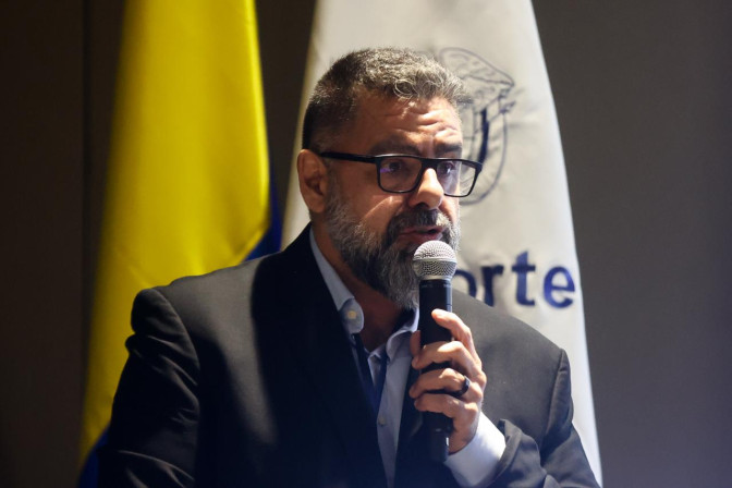 Hombre con traje oscuro, gafas y barba canosa habla al micrófono frente a la bandera de Colombia y estandarte institucional.