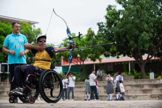 Hombre en silla de ruedas apunta con arco y flecha, acompañado por un instructor, mientras estudiantes observan al fondo.