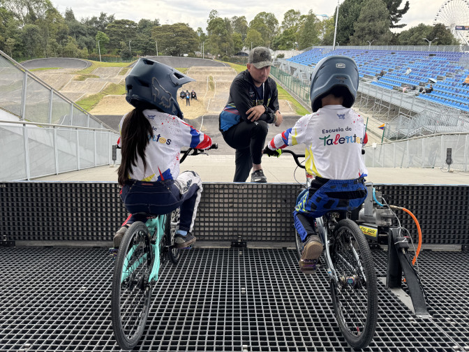 Entrenador instruye a dos jóvenes ciclistas de BMX con casco y uniforme de la Escuela de Talentos en la rampa de salida de una pista profesional.