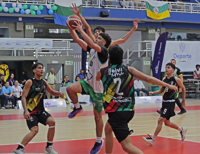 Joven jugador de baloncesto en uniforme blanco salta para anotar, rodeado de rivales con uniformes negros y verdes en cancha cubierta.