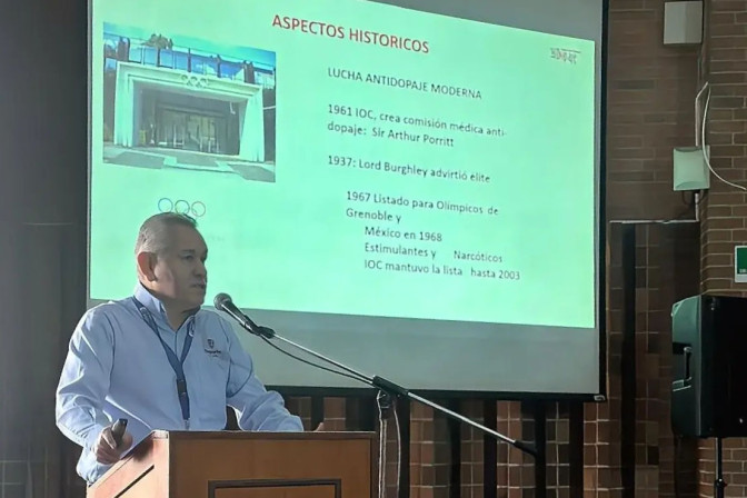 Conferencista en podio presentando diapositiva sobre historia de la lucha antidopaje moderno ante una audiencia institucional.