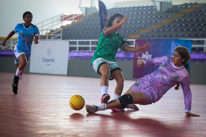 Tres jugadoras disputan un balón en partido de fútbol sala femenino en cancha cubierta durante evento deportivo.