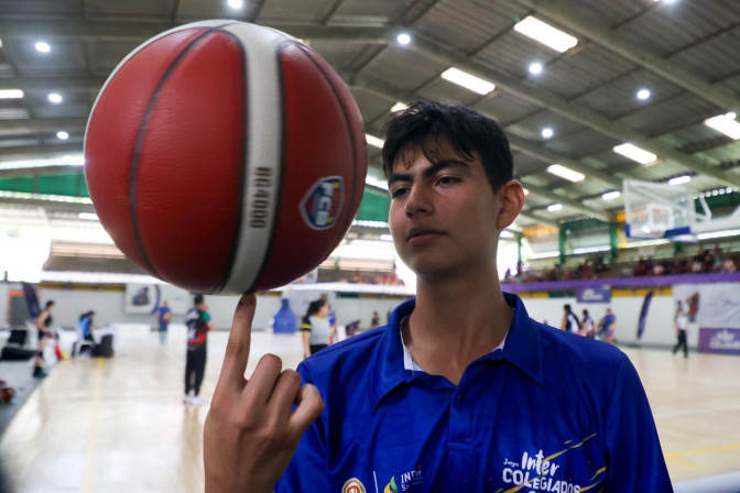 Joven con camiseta de los Juegos Intercolegiados equilibra un balón de baloncesto sobre su dedo índice en un coliseo deportivo.