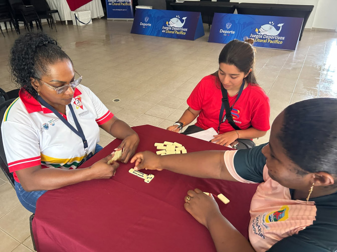 Tres mujeres juegan dominó en una mesa con mantel rojo durante los Juegos Deportivos del Litoral Pacífico.
