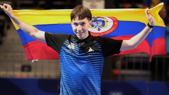 Sebastián Bedoya, tenista de mesa colombiano, celebra su medalla de oro sosteniendo la bandera de Colombia con ambas manos en alto.