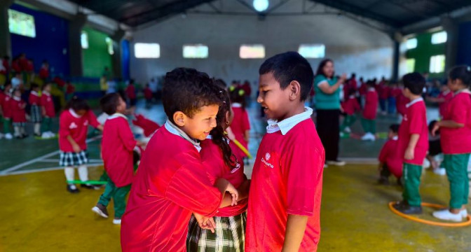 Dos niños con uniforme rojo interactúan y sonríen durante una actividad lúdica en un polideportivo escolar.