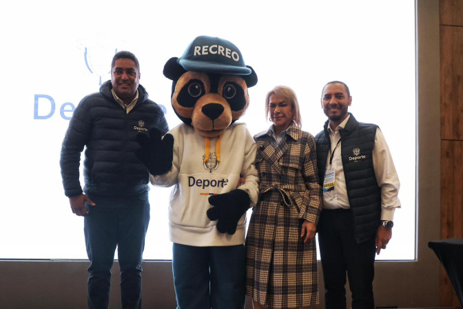 Dos hombres y una mujer con chalecos institucionales de Deporte posan junto a la mascota Recreo, un oso panda con gorra azul, frente a una pantalla de presentación.