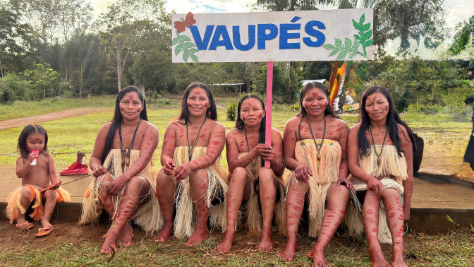Cinco mujeres indígenas del Vaupés sentadas con vestimenta y pintura corporal tradicional, sosteniendo un letrero con el nombre del departamento.