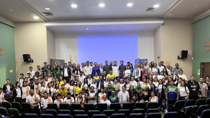 Numeroso grupo de personas posan para foto grupal en auditorio del INDER Medellín, vistiendo camisetas institucionales y deportivas.