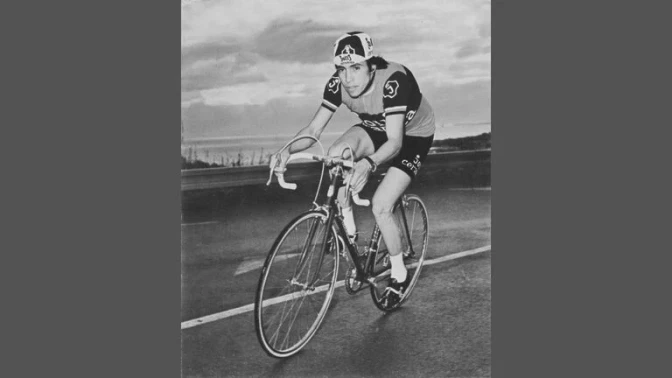 Ciclista masculino en uniforme de competencia pedalea sobre una bicicleta de ruta en una carretera abierta, fotografía en blanco y negro.