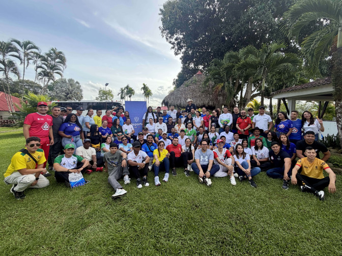 Grupo numeroso de coordinadores de campamentos juveniles posando en zona verde durante el Encuentro Nacional, con pendón institucional de Deporte al fondo.