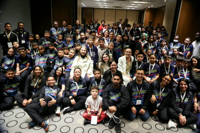 Numeroso grupo de niños y jóvenes con uniformes deportivos azules posan junto a adultos en un salón institucional