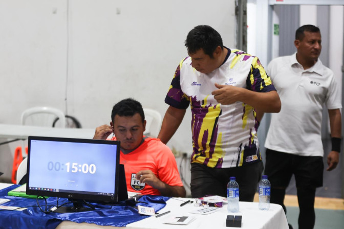 Dos hombres con camisetas deportivas operan la mesa de control de un partido de fútbol sala, con cronómetro en pantalla marcando 15 minutos.